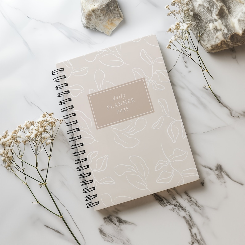 May include: A beige daily planner with a white floral pattern and the text "daily planner 2025" on the cover. The planner is lying on a white marble surface with white flowers.