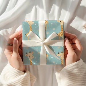 K&ouml;nnte beinhalten: Eine quadratische Geschenkbox, eingewickelt in hellblaues Papier mit einem wiederkehrenden Muster aus beigefarbenen Giraffen und wei&szlig;en G&auml;nsebl&uuml;mchen, gebunden mit einem glatten wei&szlig;en Band, das als mittige Schleife auf der Vorderseite liegt.