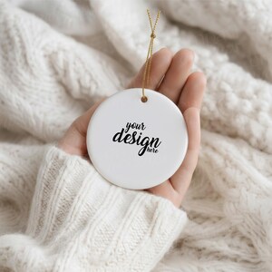 May include: A round white ceramic ornament featuring the black text your design here printed in the center. The ornament includes a small hole at the top with a thin gold string attached for hanging, providing a smooth surface for custom graphics.