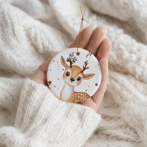May include: A round white ceramic ornament featuring a brown and white spotted fawn illustration with small antlers, decorated with gold dots and red berries, suspended by a thin gold string for hanging on a tree or display.