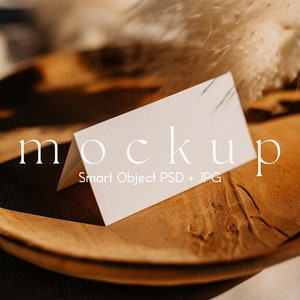 Puede incluir: Una tarjeta blanca doblada se encuentra sobre un plato de madera, con la palabra "mockup" en texto blanco. El plato tiene una veta de madera natural y está rodeado de hierba ornamental seca. El texto "Smart Object PSD + JPG" está debajo de la palabra "mockup".