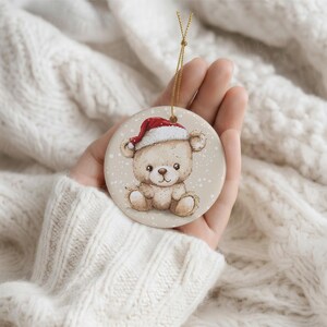 May include: A round ceramic ornament featuring a light brown teddy bear illustration wearing a red and white Santa hat. The background of the ornament is a soft beige with white speckles resembling falling snow, and it includes a gold hanging cord.