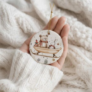 May include: A round ceramic ornament featuring a watercolor illustration of a brown bear and a white rabbit sitting together on a wooden sled. The bear wears a red scarf, and the rabbit wears a red collar. The background includes small pine sprigs.