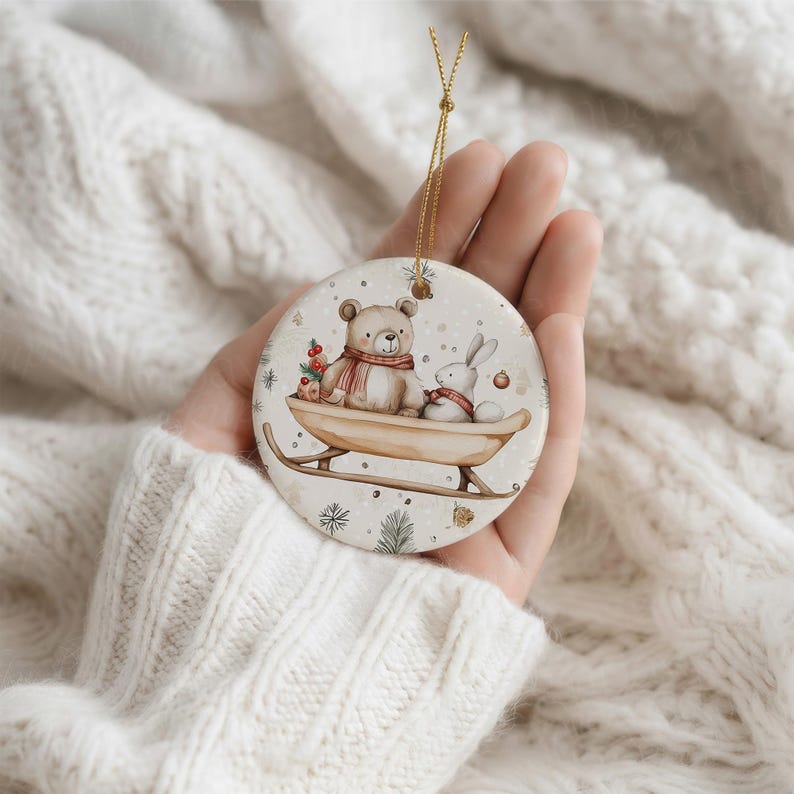 May include: A round ceramic ornament featuring a watercolor illustration of a brown bear and a white rabbit sitting together on a wooden sled. The bear wears a red scarf, and the rabbit wears a red collar. The background includes small pine sprigs.