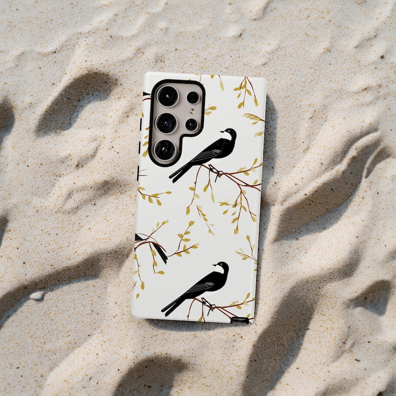 May include: A white phone case with a black camera lens and a pattern of black birds on branches with gold leaves.