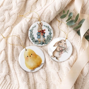 May include: Three round white ceramic ornaments with gold hanging strings. One features a brown rabbit in a blue jacket and red scarf inside a green wreath. Another shows a brown rabbit holding a floral bouquet. The third displays a fluffy yellow chick.