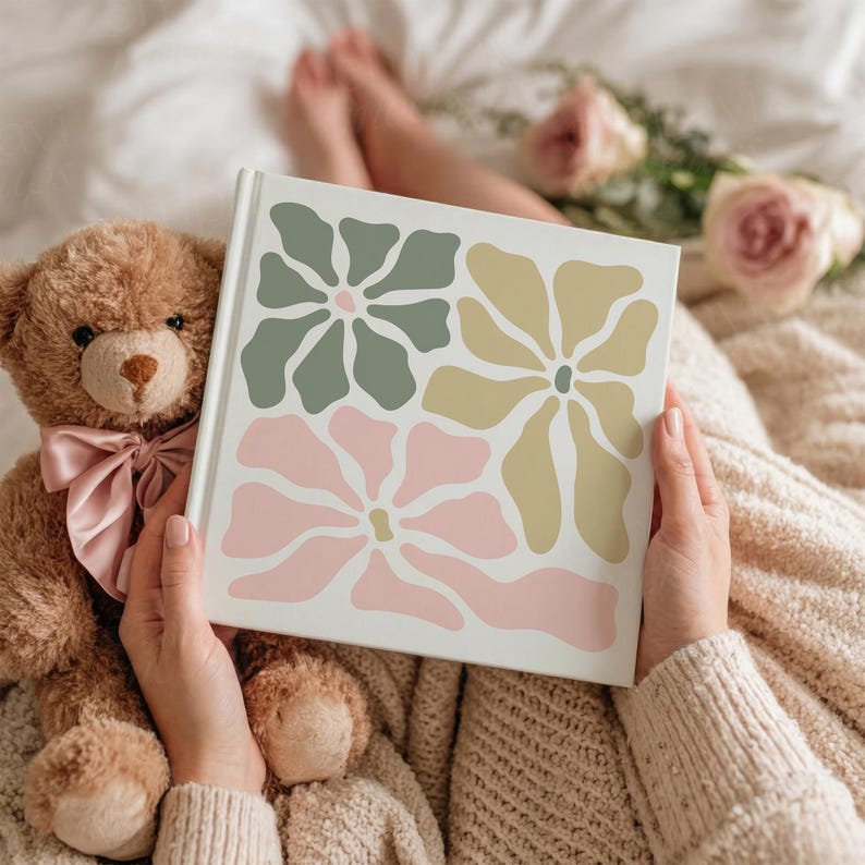 May include: A white book cover featuring a floral design in sage green, pale yellow, and pink. A teddy bear with a pink bow sits next to the book. The book is held by a person wearing a cream-colored sweater.