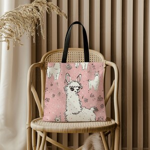 May include: A pink tote bag with a white llama design. The llama is surrounded by small pink flowers. The bag has black handles.