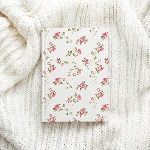May include: A hardcover journal with a white cover adorned with a repeating pattern of pink roses and green leaves. The journal is rectangular and rests on a white, knitted surface, creating a soft, floral theme.