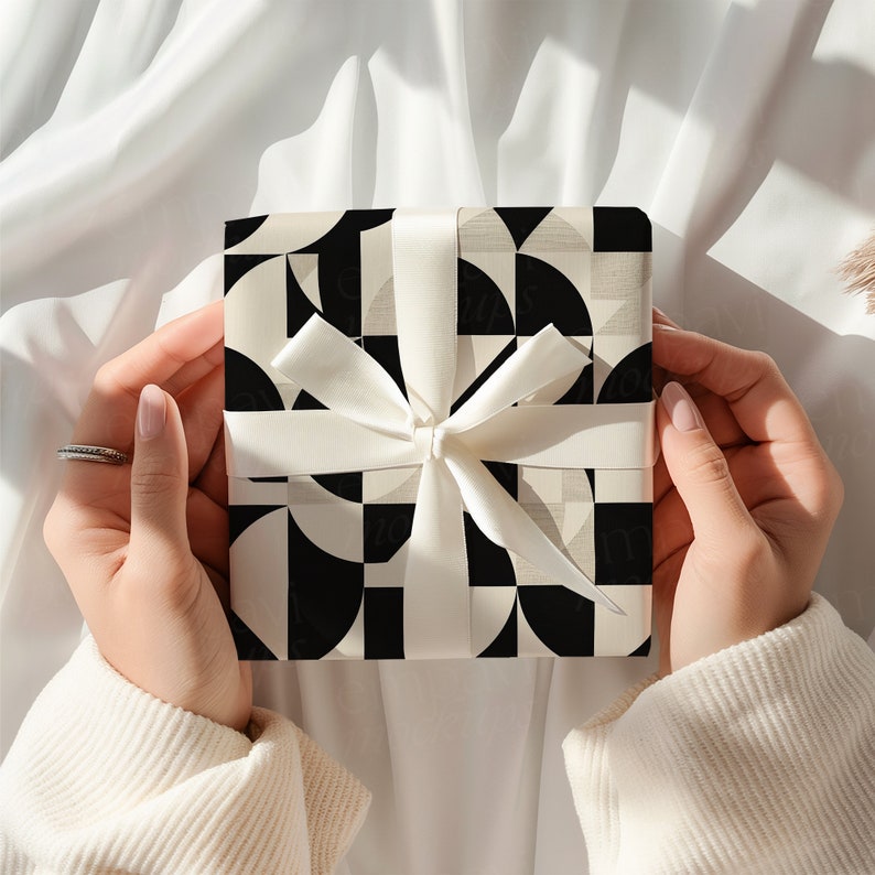 K&ouml;nnte beinhalten: Eine quadratische Geschenkbox, eingewickelt in Papier mit einem schwarz-wei&szlig;en geometrischen Muster aus sich &uuml;berschneidenden Kreisen und Quadraten. Die Box ist mit einem glatten, wei&szlig;en Satinband zu einer mittigen Schleife gebunden.