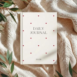 May include: A cream-colored daily journal with red heart accents scattered across the cover. The words "DAILY JOURNAL" are printed in black, with the year "2028" at the bottom. The journal is resting on a cream-colored knitted blanket.