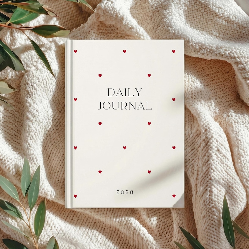 May include: A cream-colored daily journal with red heart accents scattered across the cover. The words "DAILY JOURNAL" are printed in black, with the year "2028" at the bottom. The journal is resting on a cream-colored knitted blanket.