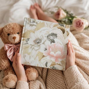 May include: A floral-patterned book with white, pink, and gold flowers is held open. A teddy bear with a pink bow sits next to the book. The background includes a soft blanket and a few roses.