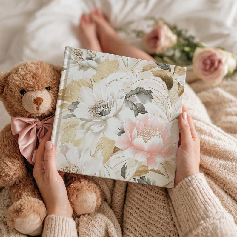 May include: A floral-patterned book with white, pink, and gold flowers is held open. A teddy bear with a pink bow sits next to the book. The background includes a soft blanket and a few roses.
