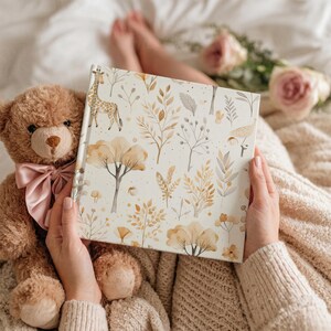 May include: A hardcover book with a botanical illustration featuring a giraffe, trees, and various plants in shades of beige, brown, and gold. The book is held in front of a teddy bear with a pink bow.