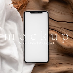 Blank black smartphone on natural wood with soft white fabric and dried leaves, creating a warm, organic setting for mobile design presentations