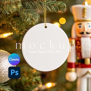 May include: A round white ceramic ornament hanging by a gold string. The ornament features the text mockup Smart Object PSD + PNG in the center. The background includes a blurred Christmas tree, a nutcracker figure, and a gold glitter ball.