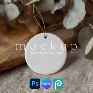 May include: A white, round ceramic ornament with a gold string for hanging. The word "mockup" is printed on the ornament. The text "Smart Object PSD + PNG" is below the word "mockup". The ornament is displayed on a wooden surface with dried leaves.