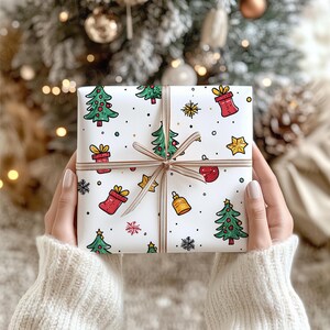 May include: A square gift box wrapped in white paper featuring a colorful pattern of green Christmas trees, red gift boxes, yellow stars, gold bells, and black snowflakes, tied with a thin, light brown ribbon centered in a cross pattern.