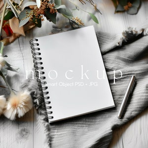 May include: A white spiral-bound notebook with a black wire coil, featuring the text mockup Smart Object PSD + JPG on the cover, accompanied by a silver metallic pen. The notebook is positioned on a grey textured fabric with frayed edges.