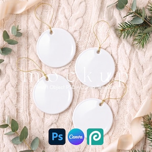 May include: Four blank white circular ceramic ornaments with gold hanging strings. The text mockup Smart Object PSD + Canva template is visible. The ornaments are arranged on a textured cream-colored knit fabric with green foliage accents.