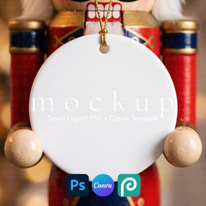 Op de afbeelding: Een rond wit keramieken ornament met een gouden ophangkoord, voorzien van de tekst mockup Smart Object PSD + Canva Template. Het ornament is geplaatst voor een rood, blauw en goudkleurige notenkrakerfiguur met wit haar.