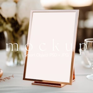 May include: A rose gold picture frame mockup on a white table with a vase of white flowers and a glass in the background. The frame is blank and ready for your design.
