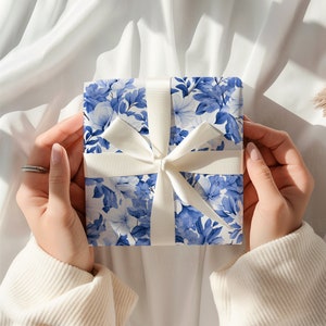 K&ouml;nnte beinhalten: Eine quadratische Geschenkbox, eingewickelt in Papier mit einem blau-wei&szlig;en Blumenmuster. Die Box ist mit einem glatten, einfarbig wei&szlig;en Band umwickelt, das zu einer mittigen Schleife gebunden ist. Das florale Design zeigt Bl&uuml;ten und Bl&auml;tter.
