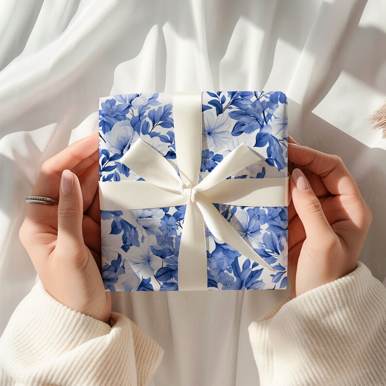K&ouml;nnte beinhalten: Eine quadratische Geschenkbox, eingewickelt in Papier mit einem blau-wei&szlig;en Blumenmuster. Die Box ist mit einem glatten, einfarbig wei&szlig;en Band umwickelt, das zu einer mittigen Schleife gebunden ist. Das florale Design zeigt Bl&uuml;ten und Bl&auml;tter.
