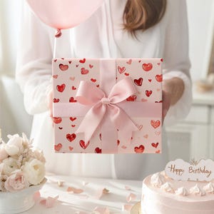 K&ouml;nnte beinhalten: Eine quadratische Geschenkbox, eingewickelt in hellrosa Papier mit einem Muster aus kleinen roten und rosa Herzen. Die Box ist mit einem breiten, glatten, hellrosa Satinband umwickelt, das zu einer gro&szlig;en Schleife auf der Vorderseite gebunden ist.