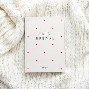 May include: A white daily journal with red heart accents. The words "DAILY JOURNAL" are printed in black, with the year "2028" at the bottom. The journal is set on a white, textured knit fabric.
