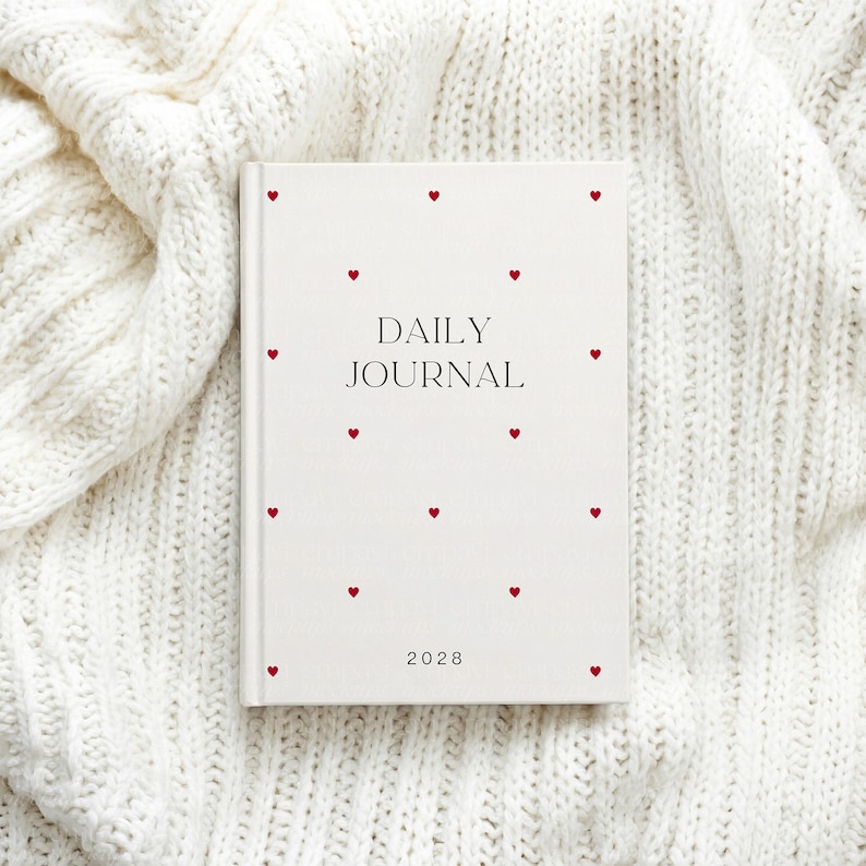 May include: A white daily journal with red heart accents. The words "DAILY JOURNAL" are printed in black, with the year "2028" at the bottom. The journal is set on a white, textured knit fabric.