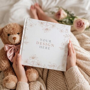 May include: A white book with the text "YOUR DESIGN HERE" in a gold color, surrounded by floral illustrations. A brown teddy bear with a pink bow sits next to the book. The scene is set on a bed with a beige blanket and flowers.