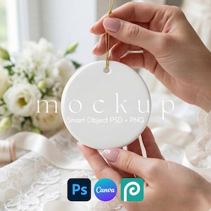 Op de afbeelding: Een witte, ronde keramische ornament met een gouden touwtje, vastgehouden door handen. Het woord "mockup" is op het ornament gedrukt. Onder het ornament staan de logo's voor Photoshop, Canva en Placeit. De achtergrond bevat witte bloemen en kant.