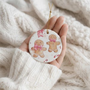 May include: A round white ceramic ornament featuring a watercolor illustration of two gingerbread cookies. One cookie wears a pink bow, and the other wears a pink bowtie. The design includes small pink accents, a candy cane, and a gold hanging string.