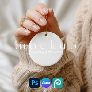 Puede incluir: Un adorno redondo de cerámica blanca con la palabra "mockup" impresa. El adorno está suspendido por un hilo dorado y sostenido por una mano. El texto "Smart Object PSD + PNG" está debajo.