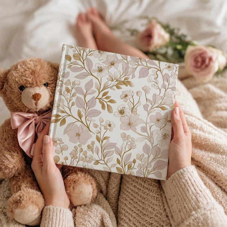 May include: A hardcover journal with a floral design in soft pink, beige, and gold tones. The journal is held open, revealing the intricate pattern of flowers and leaves. A teddy bear with a pink bow sits nearby.