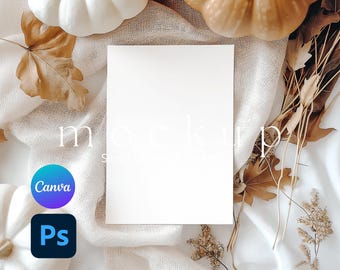 May include: A blank rectangular white card centered on a textured cream fabric, surrounded by white and tan pumpkins, dried brown autumn leaves, and thin twigs. The image features a circular Canva logo and a square Photoshop logo in the corner.