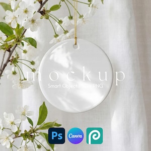 May include: A white, round ceramic ornament mockup with a gold string for hanging. The word "mockup" is printed on the ornament. The background includes white fabric and a flowering branch with white blossoms. Software icons for Photoshop, Canva, and Placeit are in the lower right.