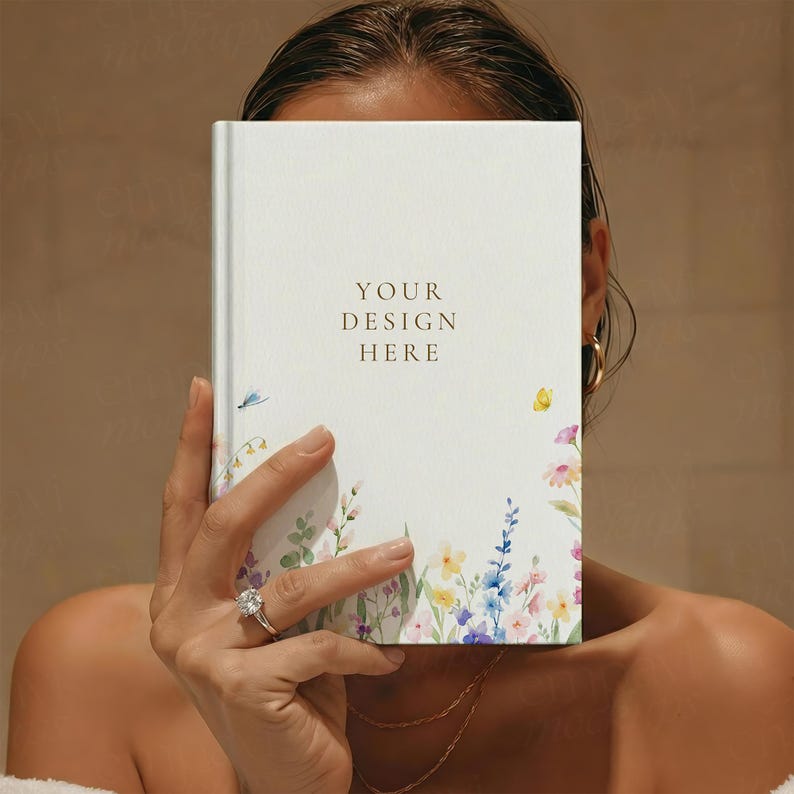 May include: A white hardback notebook featuring a delicate watercolour floral pattern along the bottom edge, including small blue, yellow, and pink flowers. The text YOUR DESIGN HERE is printed in gold in the centre of the front cover.