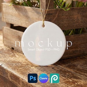 May include: A white, round ceramic ornament with a gold string for hanging. The word "mockup" is printed on the front, along with the text "Smart Object PSD + PNG". The ornament is displayed on a wooden surface with a wooden crate in the background.
