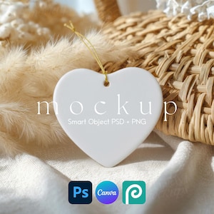 Könnte beinhalten: Ein weißer herzförmiger Ornament mit einem goldenen Faden zum Aufhängen. Das Wort "Mockup" ist auf dem Ornament aufgedruckt. Der Hintergrund umfasst einen Weidenkorb, getrocknete Pflanzen und die Logos von Photoshop, Canva und Placeit.