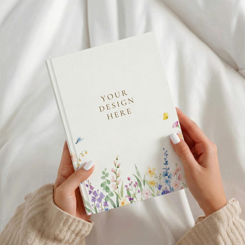 May include: A white hardback notebook featuring a delicate watercolour floral pattern along the bottom edge with various colourful wildflowers and greenery. The centre of the cover displays the gold-coloured text YOUR DESIGN HERE.