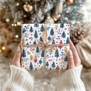 May include: A square gift box wrapped in white paper featuring a repeating pattern of dark blue evergreen trees, red berries, and yellow and blue leaves. The package is tied with a thin, light brown striped ribbon centered in a bow.