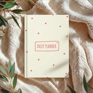 May include: A cream-colored daily planner with a repeating pattern of small red hearts. The words "DAILY PLANNER" are in a red rectangular box on the cover. The planner is resting on a cream-colored knitted blanket with green leaves in the corners.