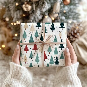 May include: A square gift box wrapped in white paper featuring a repeating pattern of stylized Christmas trees in shades of dark green, light green, red, and beige, accented with black dots and tied with a thin, metallic gold ribbon.