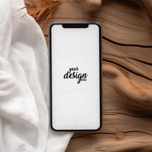 Blank black smartphone on natural wood with soft white fabric and dried leaves, creating a warm, organic setting for mobile design presentations