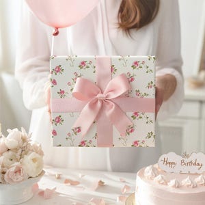 K&ouml;nnte beinhalten: Eine quadratische Geschenkbox, eingewickelt in wei&szlig;es Papier mit einem rosa Rosenmuster, gebunden mit einer breiten, glatten rosa Satinschleife. Die Box wird vor einer Person in einem wei&szlig;en Hemd gehalten, mit einem rosa Ballon &uuml;ber dem Geschenk.