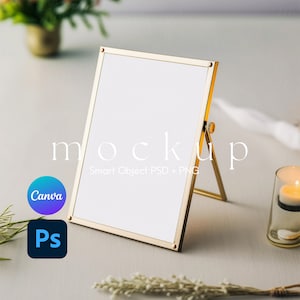 May include: A gold-colored easel-style picture frame with a white background. The frame is on a white surface with a candle and greenery in the background. The text "mockup" is written in white on the surface. The text "Smart Object PSD + PNG" is written in white below the word "mockup".