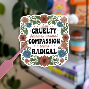 May include: A square sticker with a floral border and the text "when cruelty becomes normal compassion seems radical." The sticker features a white background, colorful flowers, and green leaves. A pink tool is in the foreground.